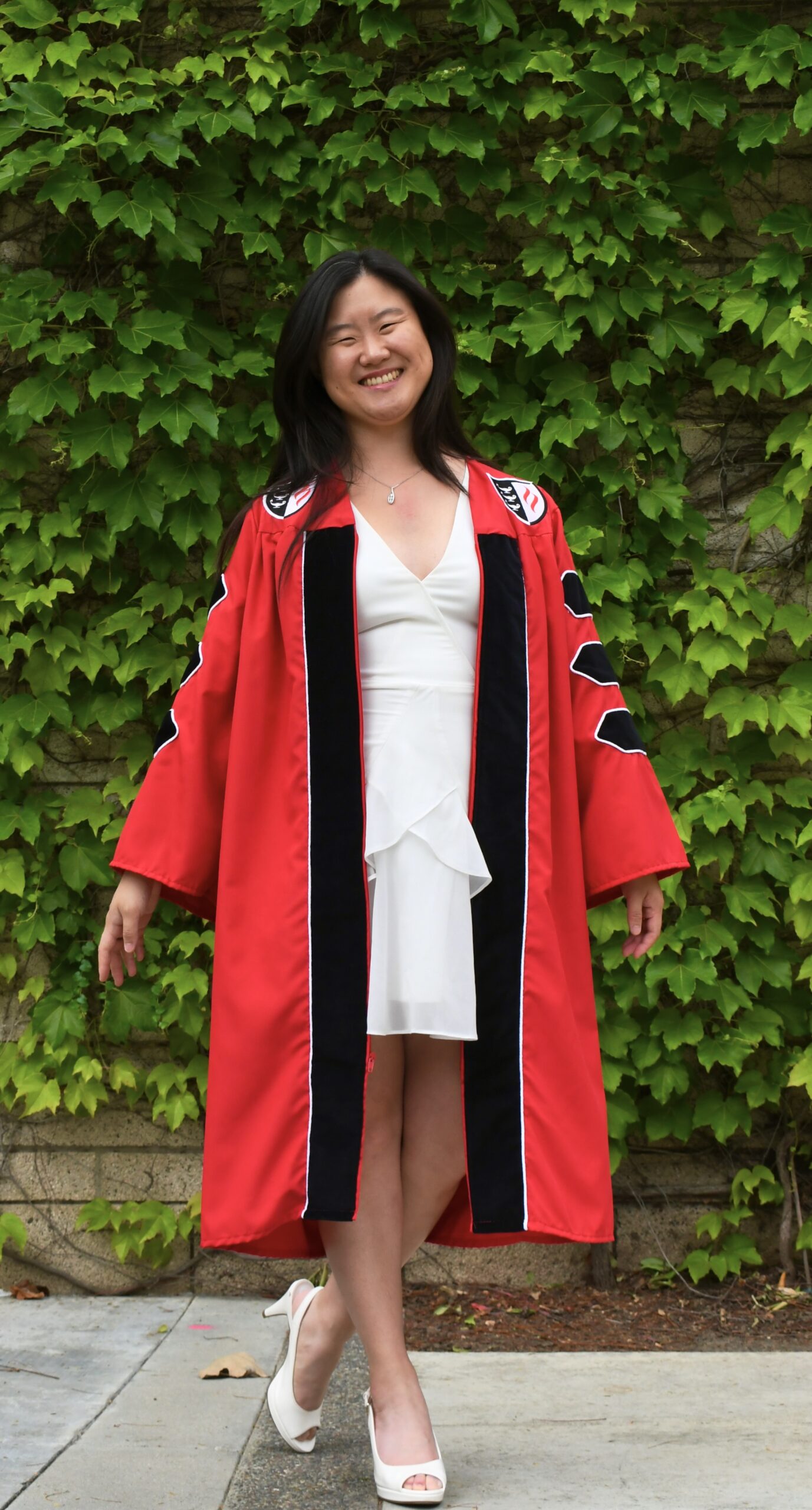Woman with black hair wearing a white dress in doctoral regalia posing in front of a wall with vines